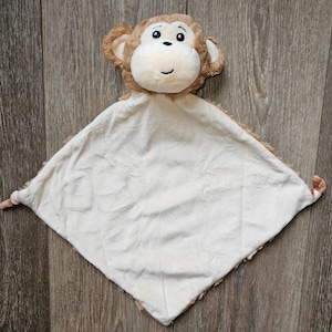 May include: A plush baby security blanket featuring a monkey head with a cream-colored face, brown ears, and a smiling expression. The blanket is cream-colored with a brown trim and is designed to comfort and soothe infants.