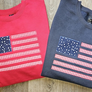 May include: Two sweatshirts, one red and one navy blue, each featuring an American flag design. The flag design includes a blue square with white stars and red and white striped accents. The sweatshirts are displayed on a wooden surface.