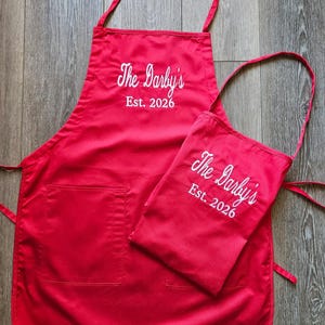 May include: Red apron and matching small apron with white embroidered text that reads "The Darby's Est. 2026". The apron has a pocket and neck and waist ties. The small apron has a neck strap.