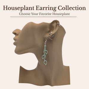 May include: A light mint green dangle earring featuring a vine-like design with three leaf-shaped cutouts, suspended from a silver-toned hook. The text Houseplant Earring Collection and Choose Your Favorite Houseplant is displayed above.