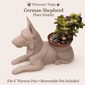 May include: A matte light brown German Shepherd plant holder featuring a sculpted dog in a lying position. The back of the dog contains a removable brown plastic pot holding a green succulent. Text reads: Winsome Wags German Shepherd Plant Holder. Fits 4" Nursery Pots • Removable Pot Included.