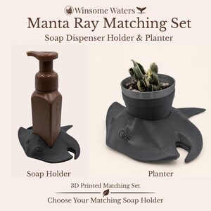 Peut inclure: Un ensemble assorti Manta Ray comprenant un porte-distributeur de savon en forme de raie manta imprimé en 3D gris foncé avec un flacon de savon marron et un pot de fleurs raie manta gris foncé avec une petite plante verte. Texte Winsome Waters.