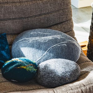 May include: A selection of stone-shaped decorative cushions in shades of grey and blue, with white detailing. The cushions are arranged on a brown, textured upholstered chair. The design creates a natural, organic look, suitable for home decor.