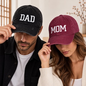 Könnte beinhalten: Eine schwarze Baseballkappe mit dem weißen gestickten Text DAD und eine kastanienbraune Baseballkappe mit dem weißen gestickten Text MOM. Beide Kappen haben ein verwaschenes Stofffinish, einen gebogenen Schirm und eine strukturierte Krone.
