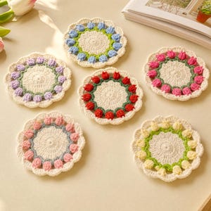 May include: A set of six round, crocheted coasters featuring a central cream-colored circle surrounded by a ring of small, three-dimensional flower buds in various colors including blue, purple, red, pink, and yellow, with green stems.