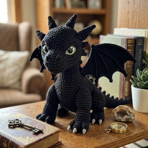 May include: A black crocheted dragon plush toy with large, light green eyes, pointed ears, and textured wings. The dragon features small white claws on its feet and is positioned on a wooden surface next to a vintage book, a metal key, and a compass.