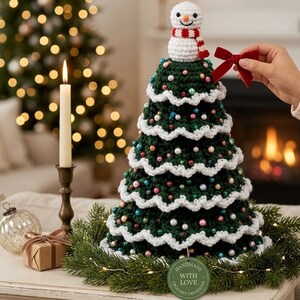 Può includere: Una decorazione per albero di Natale all'uncinetto verde scuro con bordi smerlati bianchi, perline colorate e un piccolo pupazzo di neve in cima con una sciarpa rossa e bianca. Un'etichetta verde riporta la scritta Handmade With Love Crochet Creations.