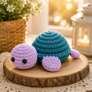 May include: A handcrafted crochet turtle toy featuring a round lavender head with two black plastic eyes, a textured teal shell, and four small lavender flippers. The stuffed animal is constructed from yarn with visible stitching throughout its surface.