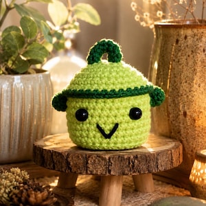May include: A light green crocheted pot with a dark green lid and handle. The pot features two black circular eyes and a black V-shaped mouth, creating a simple face. The yarn texture is visible throughout the round, lidded container design.