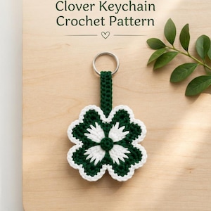 May include: A crochet clover keychain featuring a dark green four-leaf clover shape with white petal accents and a white border. The keychain includes a dark green crocheted strap attached to a silver metal ring. Text reads: CROCHET PATTERN Clover Keychain Crochet Pattern.