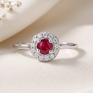 May include: A silver-toned ring featuring a round, deep red ruby center stone surrounded by a halo of small, clear, round-cut white stones. The band is smooth and polished, showcasing a classic design with a prong setting for the center stone.