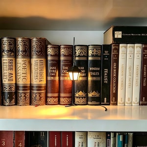 May include: A miniature black street lamp with a glowing light sits on a shelf between rows of vintage-style books. The books feature ornate gold-embossed spines with titles including Jane Eyre, Window Tales, and Feast, arranged on white shelving.