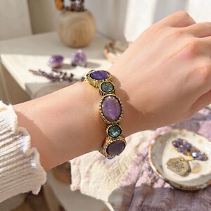 Può includere: Un bracciale con pietre cabochon ovali e rotonde alternate in tonalità viola e verde acqua, incastonate in una montatura in metallo dorato testurizzato. Le pietre sono disposte in linea, con le montature in metallo che presentano un bordo striato.