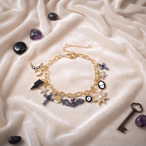May include: A gold-tone chain charm bracelet featuring various pendants including a bat, a crescent moon, a star, a cross, a lightning bolt, an eye, and a skull. The charms are accented with black enamel, clear crystals, and a central pearl.
