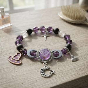 May include: A beaded bracelet featuring purple and clear faceted beads, matte black beads, and silver-toned charms. The central focal point is an oval purple stone setting flanked by two crescent moons, with a dangling crescent moon charm below.