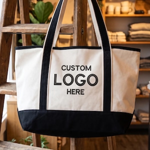 May include: A large off-white canvas tote bag with a solid black base and black fabric handles. The front of the bag features the embroidered black text CUSTOM LOGO HERE centered on the main panel, with black trim detailing along the vertical seams.
