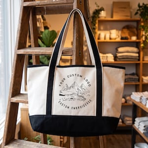 May include: A large off-white canvas tote bag with black trim and two long shoulder straps. The front features a black embroidered graphic of a mountain range and a river, with the circular text reading YOUR CUSTOM LOGO CUSTOM EMBROIDERED.