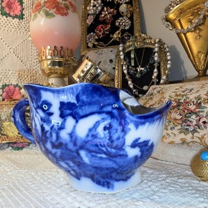 May include: A ceramic creamer pitcher featuring a blue and white transferware pattern with a scalloped rim and a curved handle. The surface displays a flowing, cloud-like floral design in deep cobalt blue against a white background.