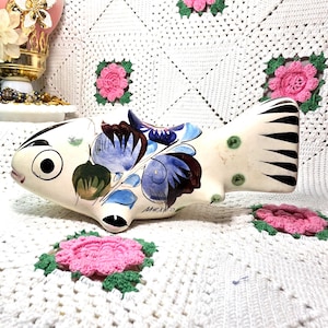 May include: A ceramic fish figurine with a cream-colored base, hand-painted with blue and dark brown floral motifs, black eye and fin details, and green dots. The word MEXICO is inscribed in black ink on the side of the fish body.