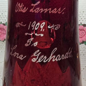 May include: A deep red glass vase featuring an etched inscription that reads Otis Lamar, 1909, To Lora Gerhardt. The surface of the glass is smooth and translucent, with a faceted base design visible at the bottom of the vessel.