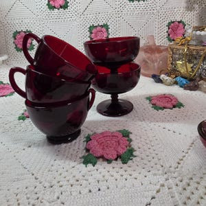 May include: A set of deep red glass drinkware consisting of three stacked cups with side handles and one stemmed glass bowl. The glassware features a smooth, translucent finish and a rounded shape, showcasing a consistent dark ruby color throughout.