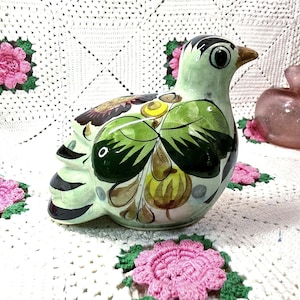 May include: A ceramic bird figurine featuring a pale green base color with hand-painted floral motifs in dark green, yellow, and brown. The bird has a pointed beak, a rounded body, and black painted details on the head and tail feathers.