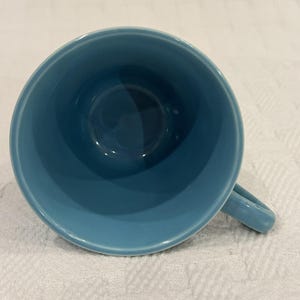 Puede incluir: Una vista desde arriba de una taza de cerámica de color azul claro sólido con un acabado suave y brillante. La taza presenta una abertura circular y un asa curva única que se extiende desde el lado, mostrando un color uniforme en el interior.