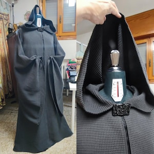 Peut inclure: Une robe à capuche gris foncé avec une texture gaufrée. La robe présente un design long et fluide avec de larges manches et un fermoir décoratif au niveau du col. La capuche est grande et drapée, et la robe est présentée sur un mannequin.