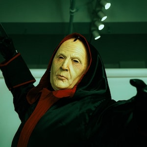 May include: A person wearing a black hooded robe with a red lining, holding a knife in their right hand. The person is wearing a white mask with a stern expression.