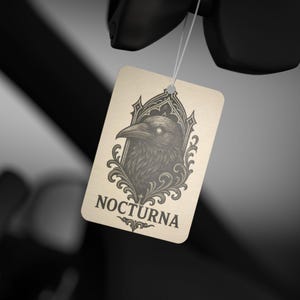 May include: A rectangular tag with a detailed illustration of a raven's head within an ornate frame. The tag has the word "NOCTURNA" printed in a serif font. The tag is attached to a black object with a white string.
