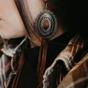 May include: Silver-toned oval earrings with a decorative scalloped edge and a central brown inlay. Long brown leather fringe hangs from the bottom of the earrings. The earrings are worn with a black top and a plaid shirt.