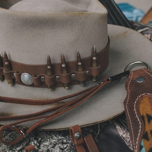 Pode incluir: Um chapéu de cowboy cinza claro com uma faixa de couro marrom adornada com estojos de balas. Uma alça de couro combinando e uma peça decorativa de couro com um padrão também estão presentes.