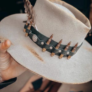 May include: A light beige cowboy hat with a distressed brim. The hat features a black leather band adorned with brass bullet casings and a silver emblem. A feather is attached to the side of the hat, adding a decorative touch.