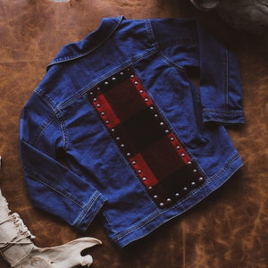 May include: A blue denim jacket with a decorative back panel featuring a red and black plaid design. The plaid is accented with silver-coloured studs. The jacket is laid flat on a brown leather-like surface.