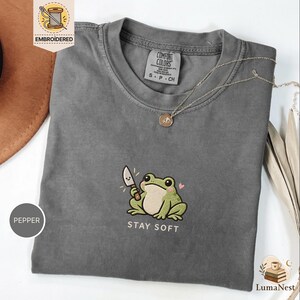 May include: A charcoal grey t-shirt featuring an embroidered graphic of a green frog holding a small knife with a smiling face. Below the frog, the text reads STAY SOFT in white capital letters. The shirt has a crew neckline and a size S label.