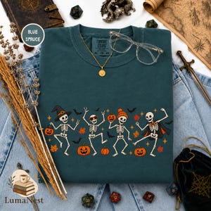 May include: A blue spruce crewneck t-shirt featuring a graphic of four dancing skeletons with Halloween elements including pumpkins, bats, and autumn leaves. The skeletons wear accessories like a witch hat, sunglasses, a beanie, and a cape.