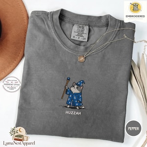 以下が含まれることがあります： チャコールグレーの半袖Tシャツ。青いローブと帽子を身につけ杖を持った魔法使いの猫の刺繍が施されています。猫の下にはHUZZAHという文字が刺繍されています。TシャツにはComfort Colorsのブランドラベルが付いています。