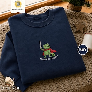 May include: A navy blue crewneck sweatshirt featuring an embroidered graphic of a green chameleon wearing a red cape and holding a silver sword. Below the chameleon, the text reads Master Of Disguise in white lettering. The fabric has a smooth texture.