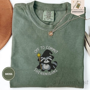 May include: A moss green t-shirt featuring an embroidered graphic of a raccoon wearing a black witch hat and cape, holding a yellow star wand. The text reads OFF TO COMMIT SHENANIGANS. The shirt has a Comfort Colors label at the inner neckline.