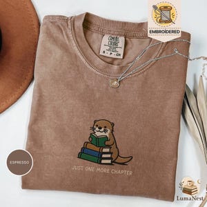 Op de afbeelding: Een espresso-kleurig t-shirt met een geborduurde afbeelding van een otter die een boek leest terwijl deze op een stapel van drie boeken zit. Onder de otter staat de tekst JUST ONE MORE CHAPTER. Het shirt heeft een Comfort Colors neklabel.