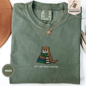 May include: A moss green t-shirt featuring an embroidered graphic of a brown otter sitting and reading a green book atop a stack of three books. Below the otter, the text reads "JUST ONE MORE CHAPTER" in white. The shirt has a crew neckline.