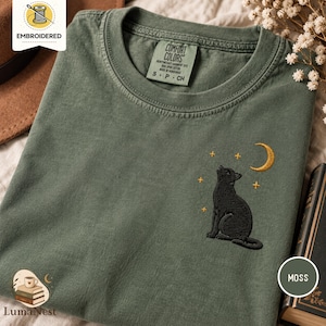 Pode incluir: Uma t-shirt de gola redonda verde musgo com um design bordado de um gato preto sentado a olhar para cima para uma lua crescente dourada e pequenas estrelas. A etiqueta Comfort Colors no interior da gola indica o tamanho small.