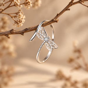 May include: A silver-toned open ring featuring a dragonfly motif with cut-out wing details and a single small clear stone set at the center of the dragonfly body. The ring is resting on a thin, brown, leafless branch.