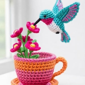 Puede incluir: Un colibrí de ganchillo hecho con hilo turquesa, rosa y blanco que flota sobre un grupo de flores rosas con centros amarillos. Las flores están plantadas en una pequeña taza de té a rayas naranjas y rosas sobre un platillo a juego.