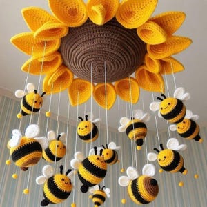 May include: A hanging mobile featuring a large, crocheted sunflower with a brown centre and yellow petals. Suspended from the sunflower by white strings are multiple small, crocheted bumblebees with yellow and black striped bodies and white wings.