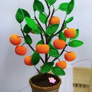 Peut inclure: Un oranger au crochet dans un pot marron. L'arbre présente des branches marron foncé, des feuilles texturées vert vif et plusieurs fruits ronds orange vif. Une petite fleur rose est fixée sur la terre marron foncé dans le pot.
