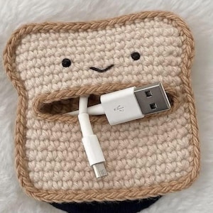 Puede incluir: Un organizador de cables de ganchillo beige con forma de rebanada de pan y borde de corteza marrón oscuro. La cara tiene dos pequeños ojos negros bordados y una boca curva. Un cable de carga USB blanco atraviesa la abertura central.