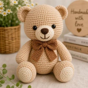 Puede incluir: Un oso de peluche tejido a ganchillo en color beige claro con una nariz bordada en marrón oscuro, ojos de plástico negro y un hocico texturizado. El oso lleva una pajarita tejida en marrón oscuro y parches circulares claros en los pies.