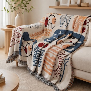 Puede incluir: Una manta tejida con base color crema y un patrón gráfico repetitivo en azul, naranja y rojo. El diseño incluye gatos, peces, setas y motivos florales estilizados, rematado con un borde de flecos en la parte inferior de la pieza.
