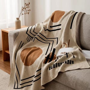 Könnte beinhalten: Eine Strickdecke mit cremefarbener Basis und einem Muster aus braunen Kreisen sowie schwarzen geometrischen Linien. Die Decke liegt über einem Sofa und zeigt ein strukturiertes Gewebe mit einem durchgehenden abstrakten Design.